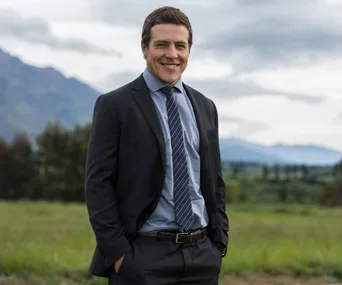 A man in a suit smiles outdoors with a mountain landscape in the background.