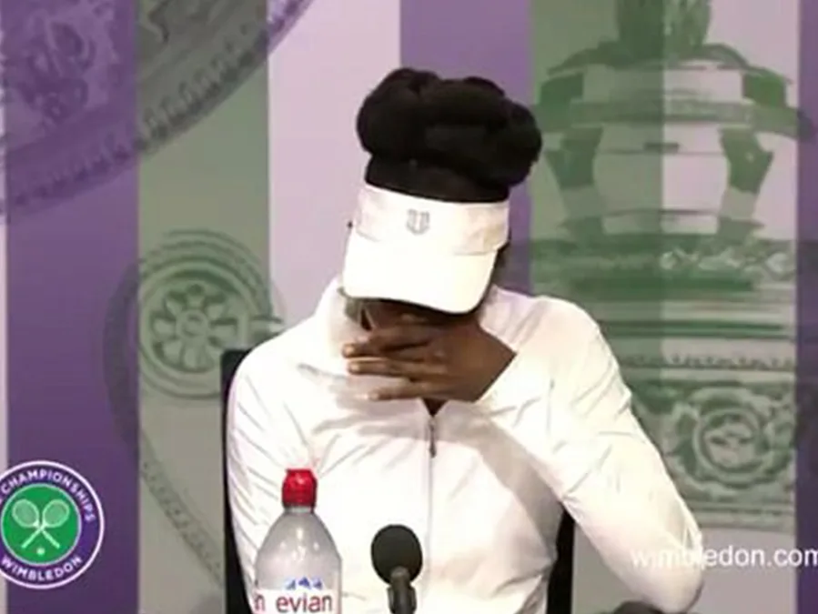 Tennis player at a Wimbledon press conference, emotionally wiping face with hand under white cap and jacket.
