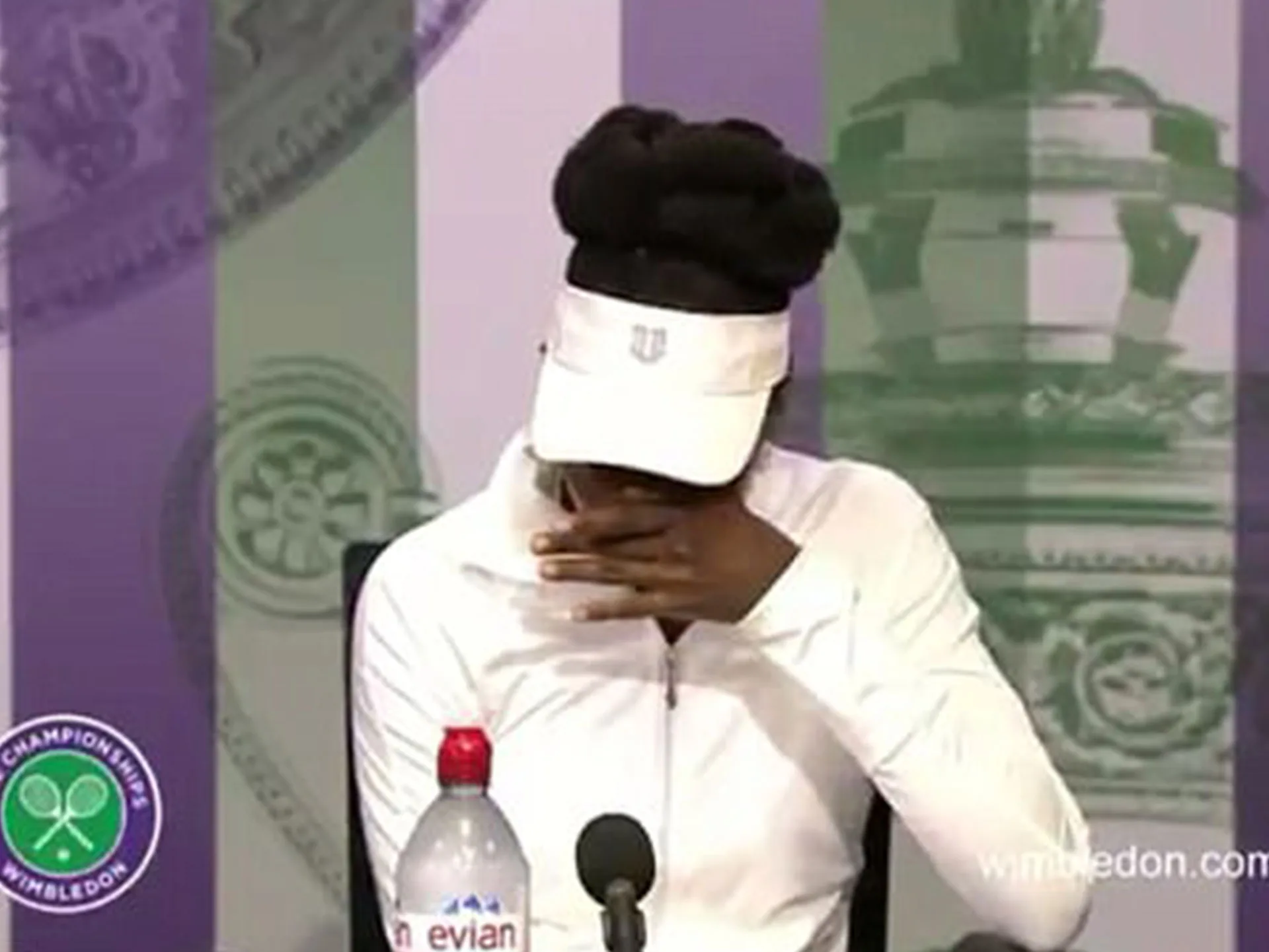Tennis player at a Wimbledon press conference, emotionally wiping face with hand under white cap and jacket.
