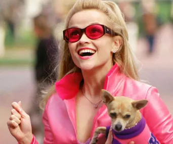 Blonde woman in pink outfit holding a small dog, wearing red sunglasses, smiling outdoors.