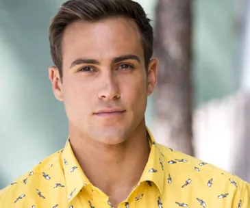 Matt Wilson as Aaron Brennan from Neighbours, wearing a yellow shirt with bird designs, outdoor background.
