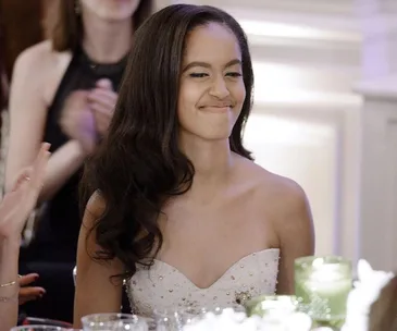 Young woman in an elegant dress, smiling at an event with people in the background.