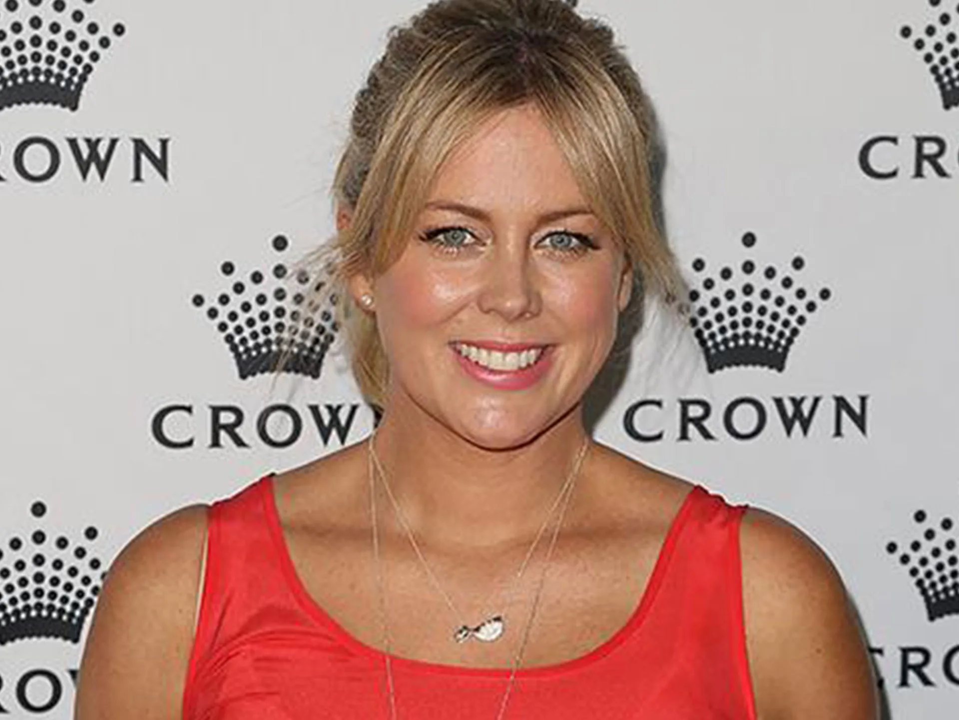 A person smiling in front of a Crown backdrop, wearing a red sleeveless top and a necklace.