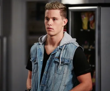 Young man in denim vest stands in front of vending machine, looking serious.