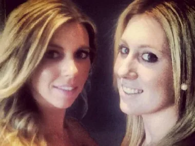 Two women smiling, with long blonde hair, posing closely together against a dark background.