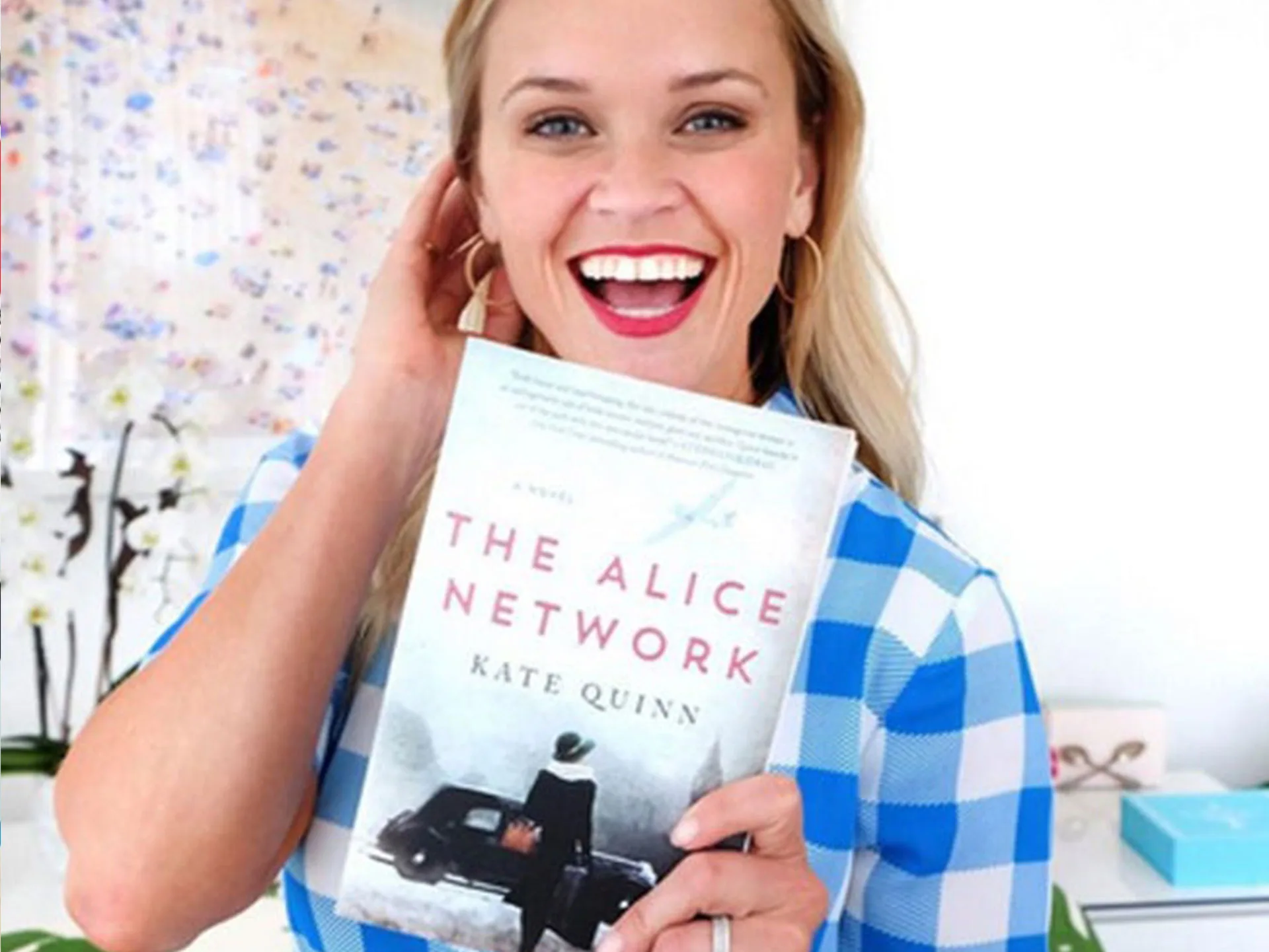Woman in a blue checkered shirt holding "The Alice Network" book by Kate Quinn, smiling.