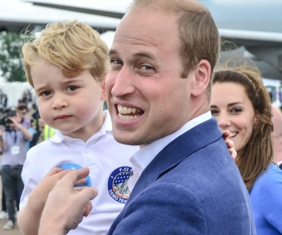 Man smiles holding a young boy, with a woman in the background, suggesting a family moment outdoors.