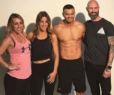 Group photo of four smiling people posing together, one shirtless male with defined abs.
