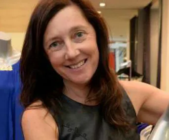A smiling woman with dark hair, wearing a sleeveless top, stands indoors near clothing displays.