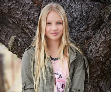 Young girl with long blonde hair wearing a green jacket, standing in front of a large tree trunk.
