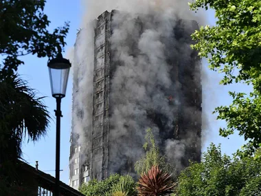 A charred high-rise tower emitting smoke, surrounded by trees, under a clear sky.