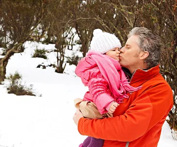 Father kissing daughter in snowy forest, both in warm clothes; she wears a pink jacket and white hat.
