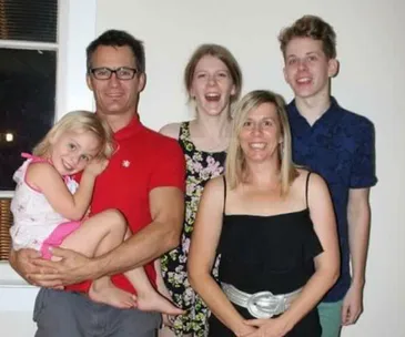 A family of five smiling indoors; man holding young girl, two teenagers, and a woman.