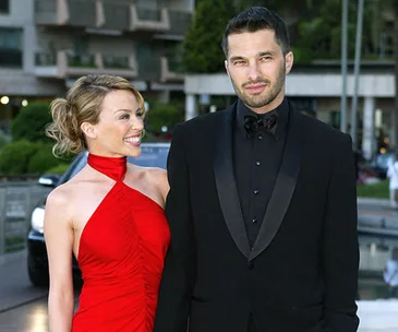 Man and woman on red carpet, woman in red dress smiling at man in black suit, outdoor event backdrop.
