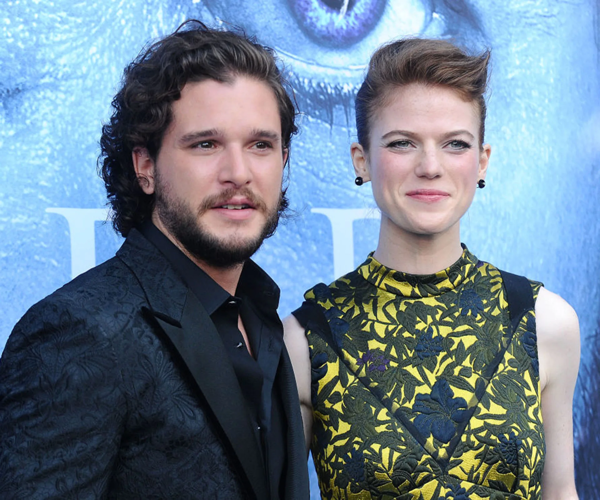 A couple poses at an event, the man in a dark suit and the woman in a yellow floral dress against a blue background.