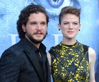 A couple poses at an event, the man in a dark suit and the woman in a yellow floral dress against a blue background.