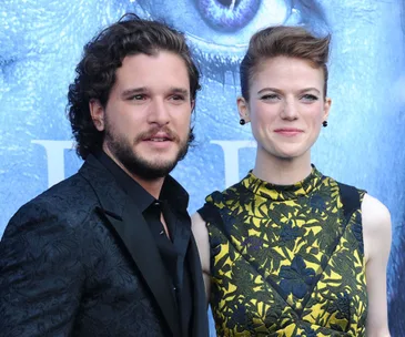 A couple poses at an event, the man in a dark suit and the woman in a yellow floral dress against a blue background.