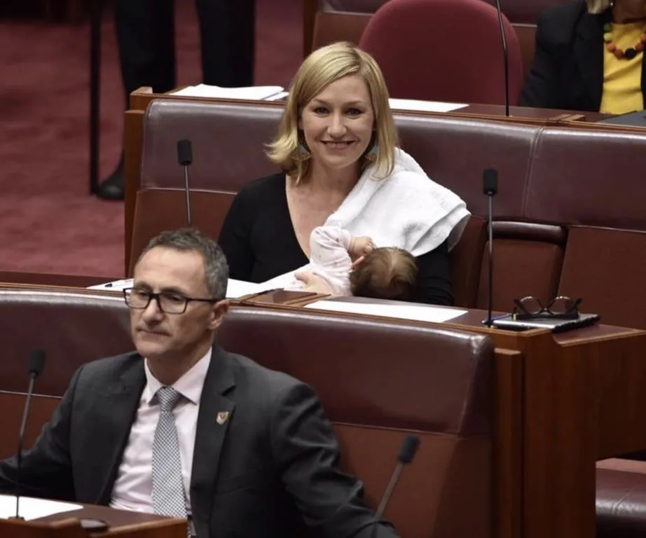 A woman in a legislative chamber is smiling while breastfeeding a baby, with a man in a suit sitting in front of her.