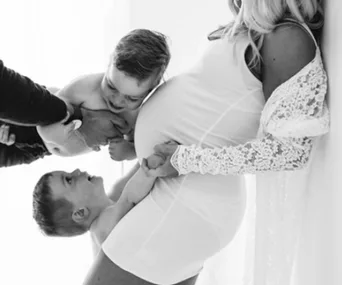 Pregnant woman in white dress, lying down with two young children holding hands and another being lifted above her.