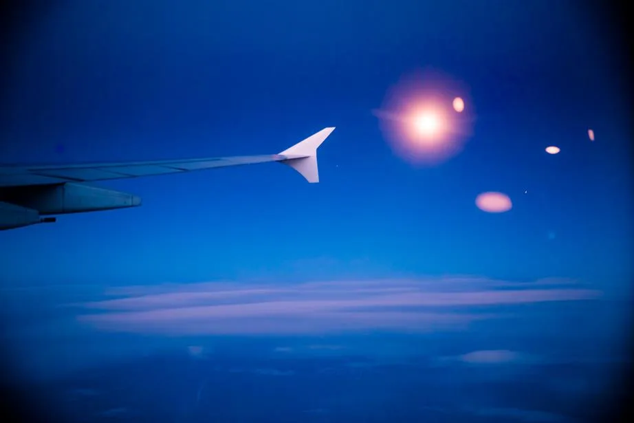 Airplane wing flying above clouds at sunset, with lens flares against a deep blue sky.