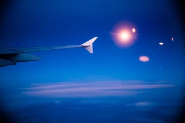 Airplane wing flying above clouds at sunset, with lens flares against a deep blue sky.