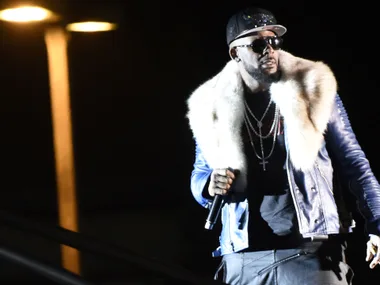 A performer on stage wearing a fur-collared jacket, sunglasses, and holding a microphone.