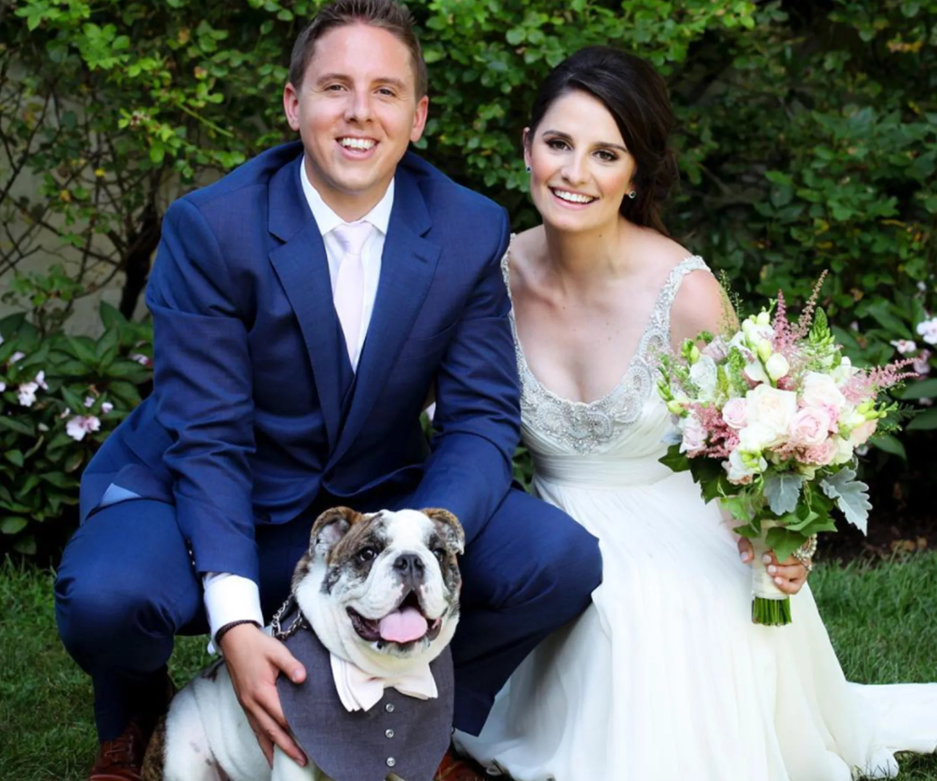 Bride and groom kneeling beside a dressed bulldog outdoors, bride holding a flower bouquet, all smiling.