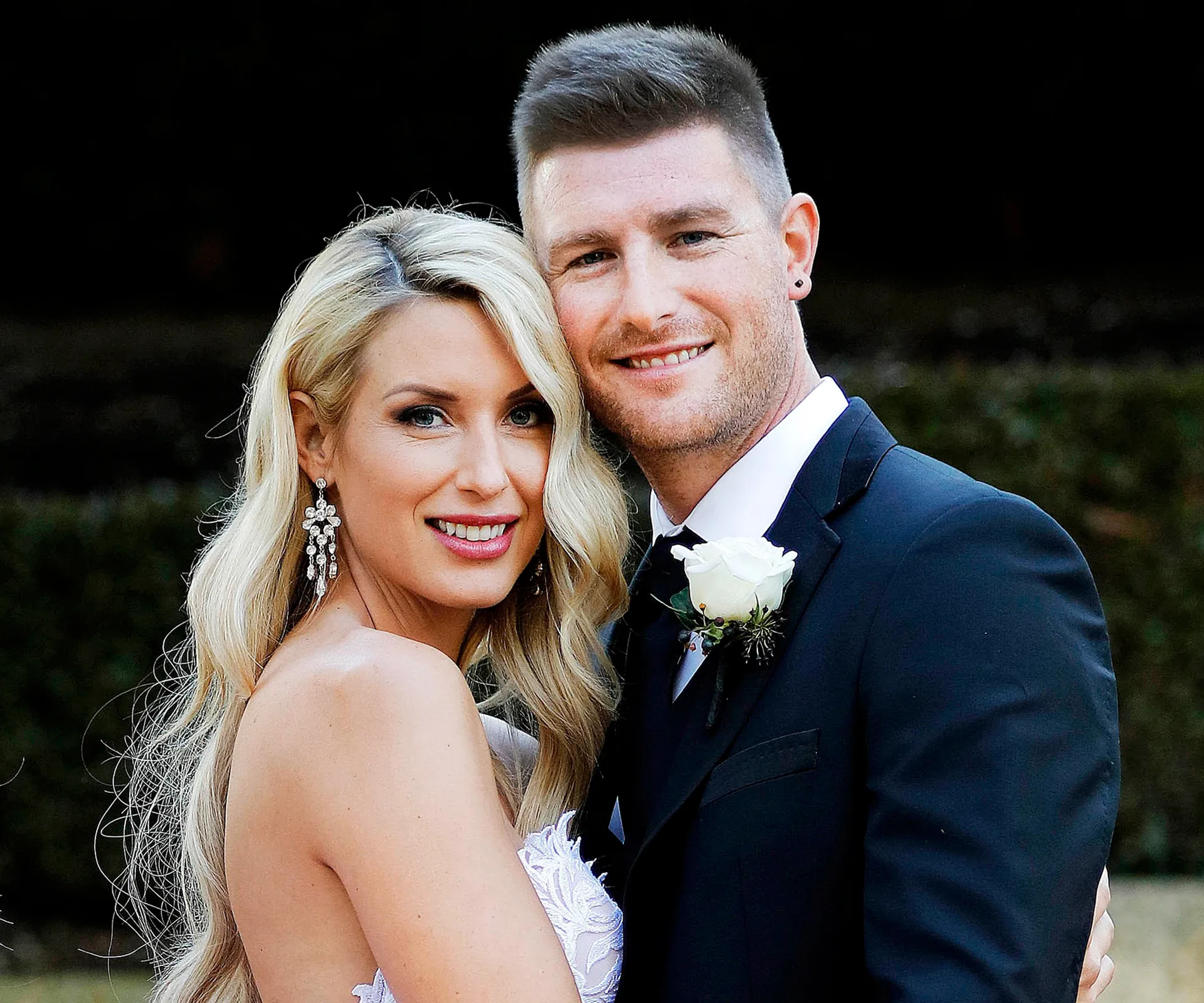 A couple dressed in wedding attire, embracing and smiling at the camera.