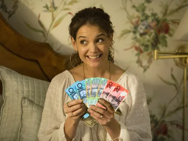 Woman smiling and holding colorful currency notes in a cozy room with floral wallpaper and a quilted bedspread.