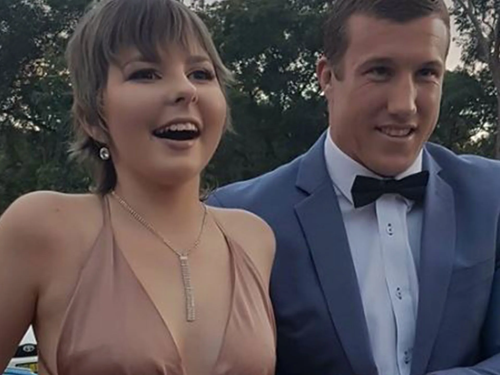 Two people smiling, one in a formal suit and the other in a dress, standing outdoors with trees in the background.