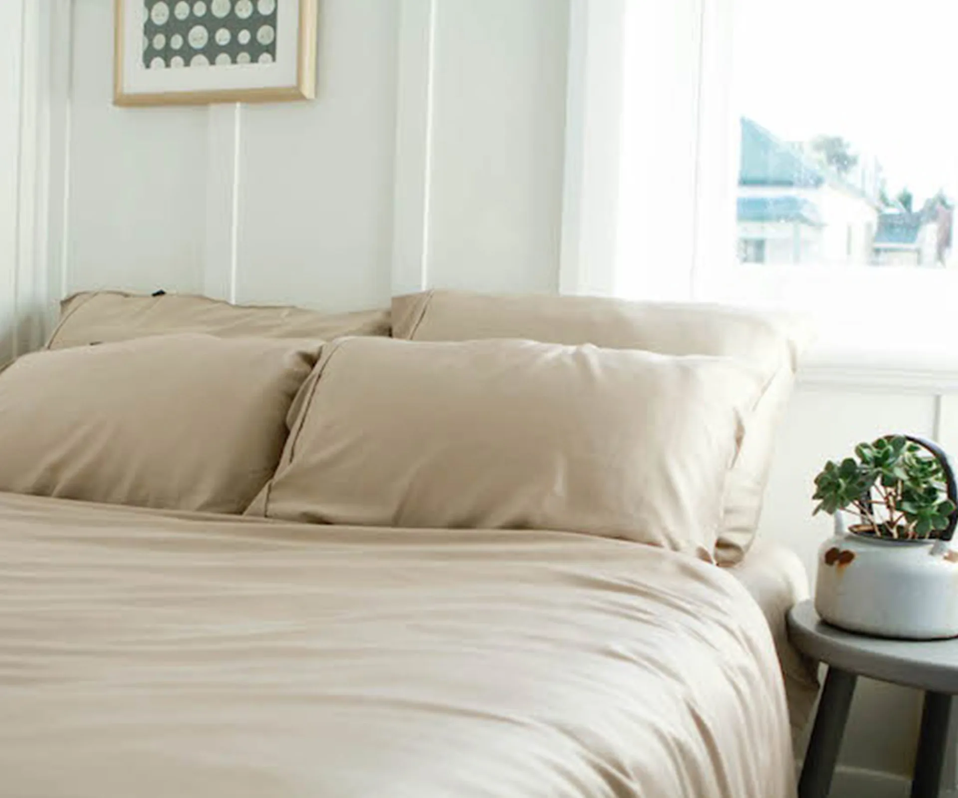 Beige bed sheets on a neatly made bed with a potted plant and framed artwork in a bright room.