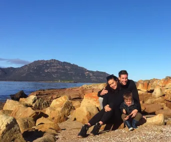 A couple and a child sit on rocky shore with mountain in background, under a clear blue sky.