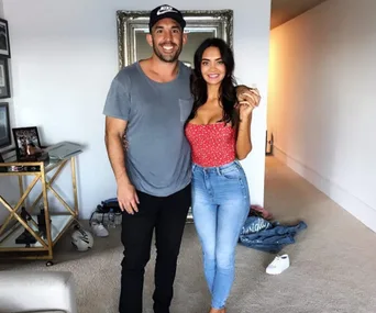 Two people smiling and posing indoors, with casual clothing and a room with mirrored furniture in the background.