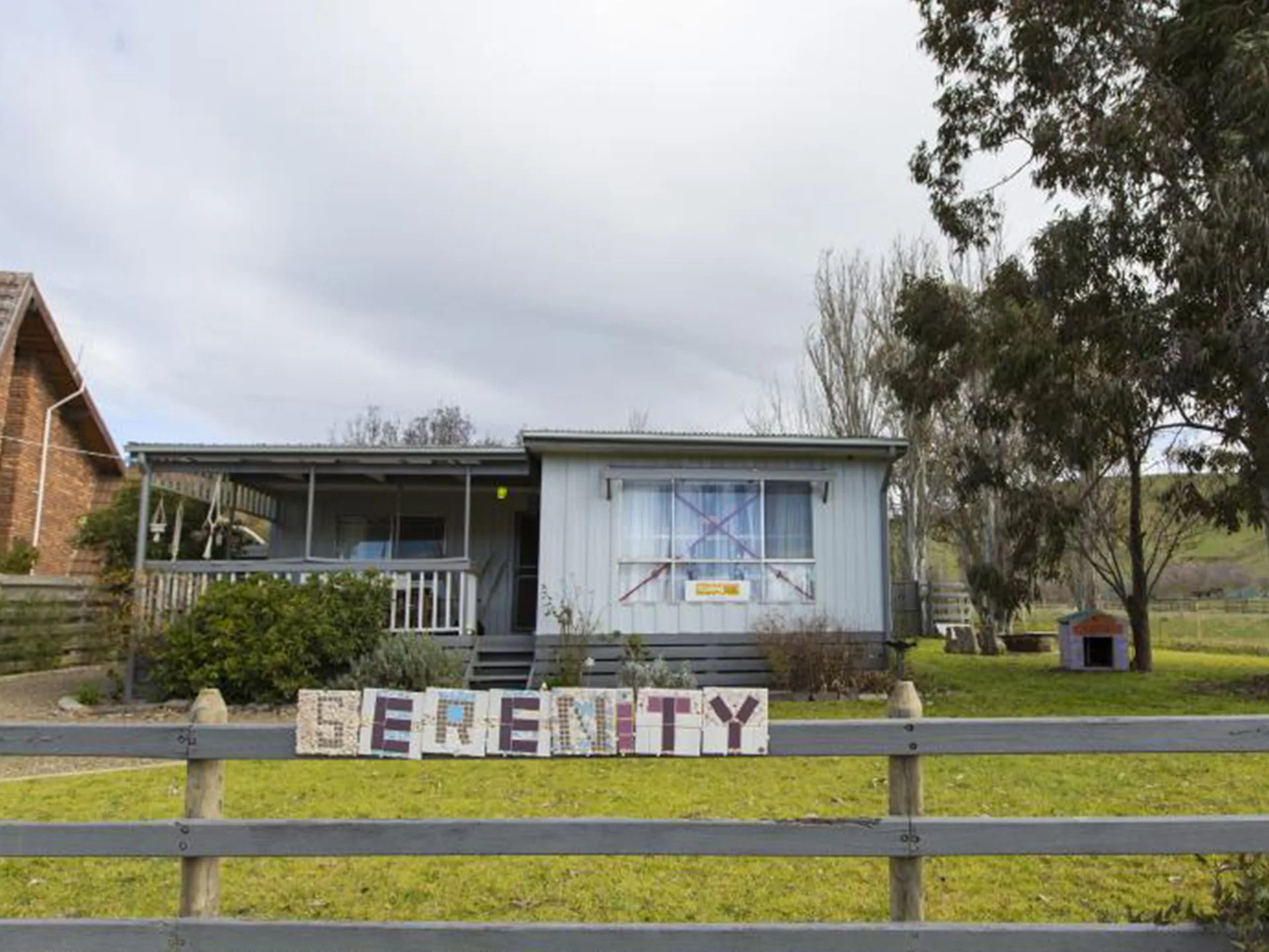 House from "The Castle" movie with "Serenity" sign in front, located on a grassy plot with trees in the background.