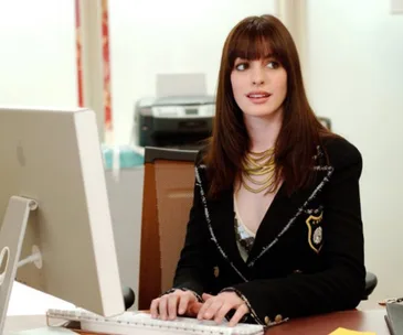 A person with long brown hair sits at a desk, typing on a keyboard, looking to the side in an office setting.