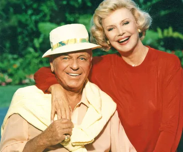 An elderly couple smiling, the man wearing a hat and sweater, the woman in a red top with greenery in the background.