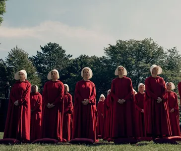Handmaids in red robes and white bonnets stand in formation on green grass, facing forward with trees in the background.
