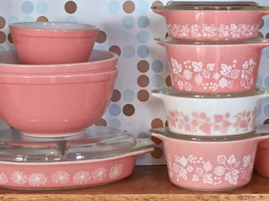 Vintage pink and white Pyrex bowls and dishes with floral patterns stacked on wooden shelf.