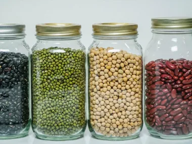 Four mason jars filled with different beans: black, green, beige, and red, aligned on a white surface.