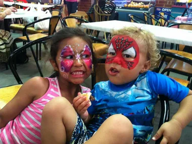 Two children with colorful face paint sit in a cafe: one has a butterfly design, the other has a Spider-Man mask.