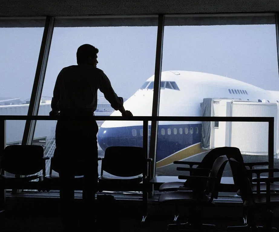 Silhouette of a person looking at a plane through terminal window, chairs and glass wall visible.