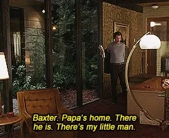 Man entering with newspaper, speaking to small dog named Baxter in a cozy living room setting. Subtitles read: "Baxter, Papa's home..."