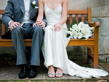Bride and groom seated on a wooden bench, holding hands with a bouquet of white lilies beside them.