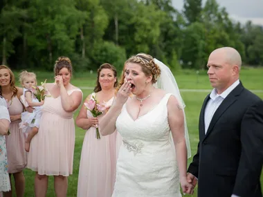 Bride in white gown reacts emotionally, surrounded by bridesmaids in pink dresses, holding a man's hand outdoors.