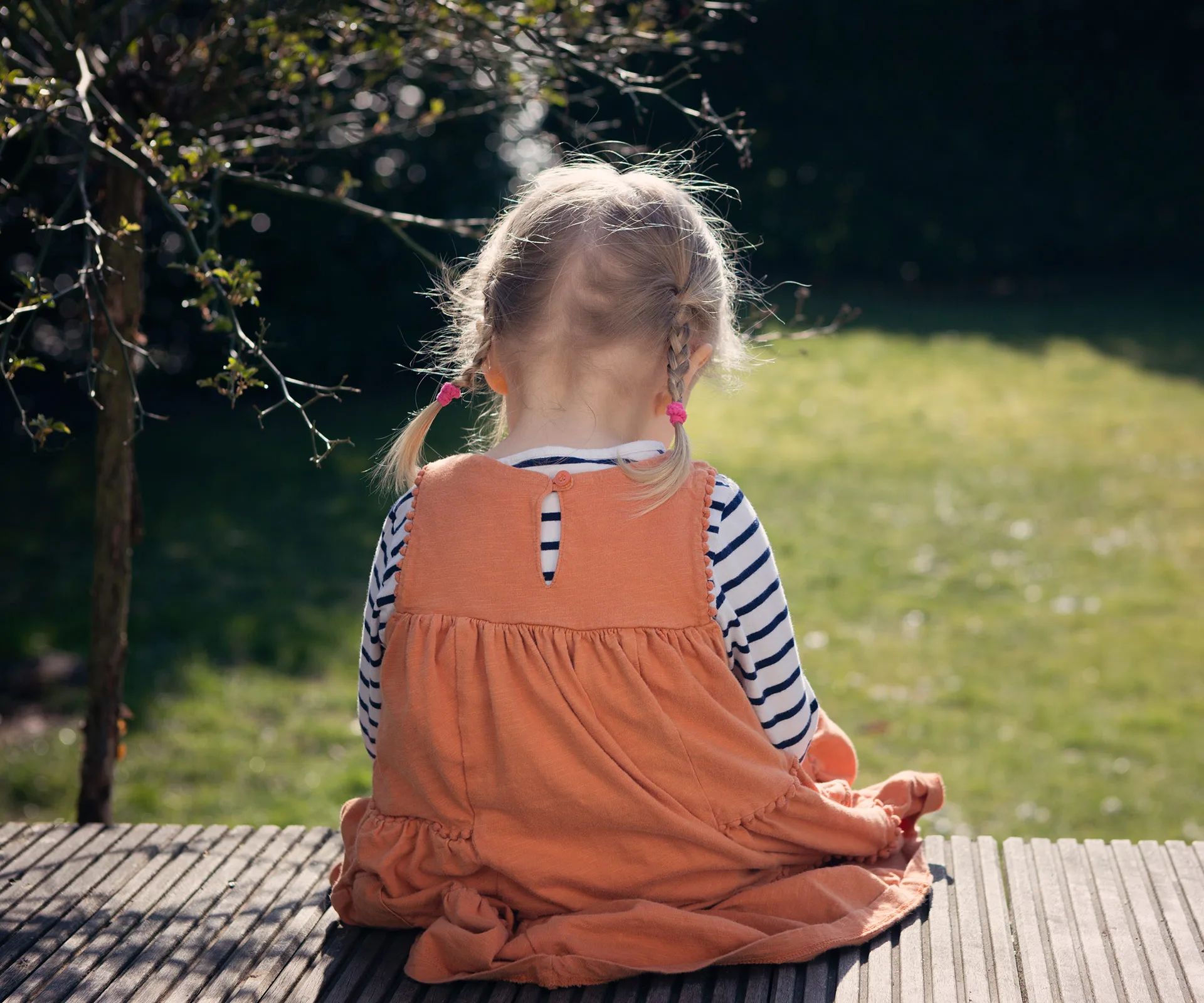 A young girl with pigtails sits on a wooden deck, wearing an orange dress over a striped shirt, facing away from the camera.