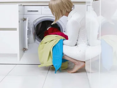 A woman crouching in front of a washing machine, holding colorful clothing items.