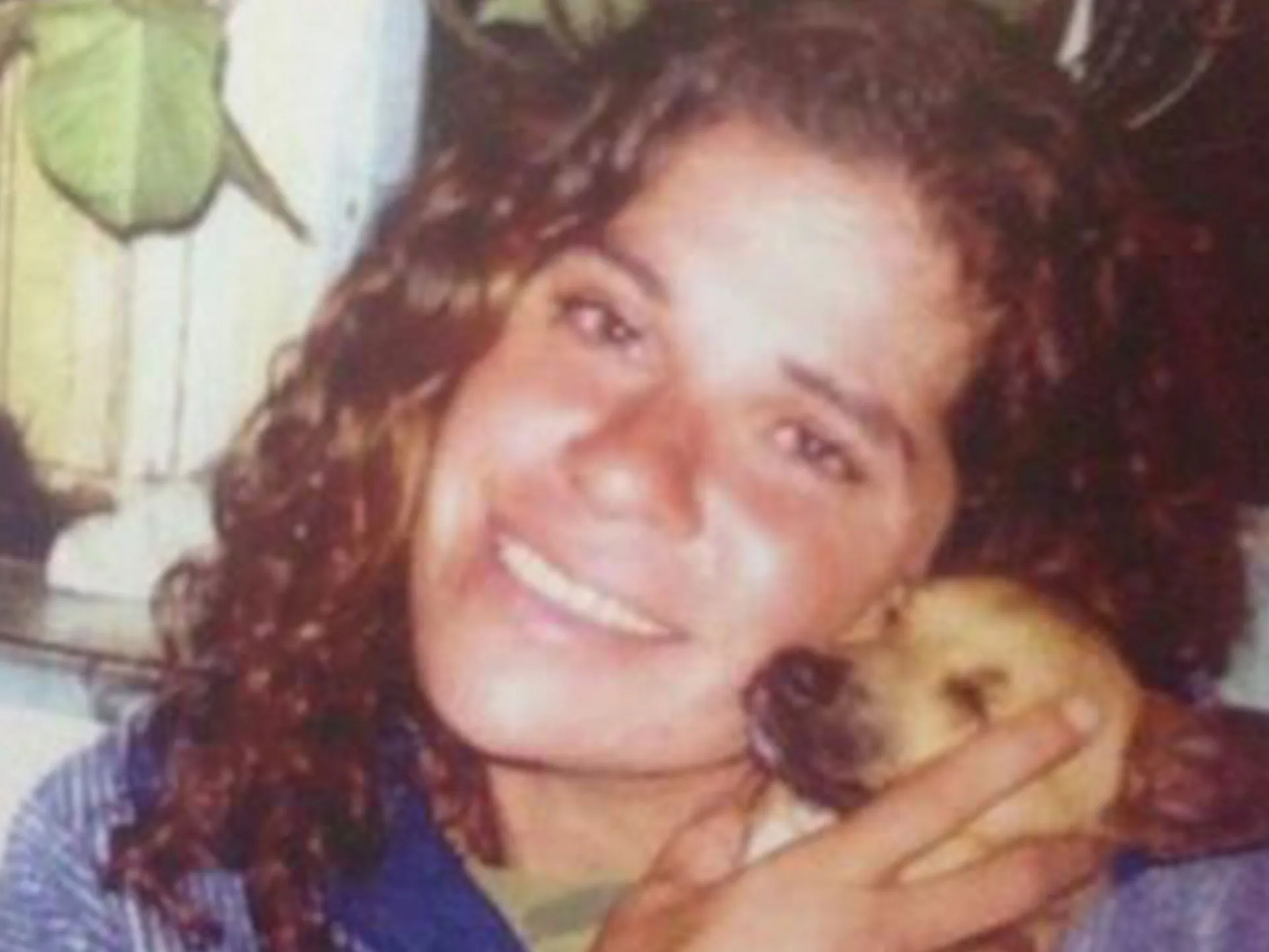 Woman smiling and hugging a small dog, with plants in the background.