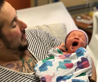 A bearded person wearing a striped shirt holds a yawning newborn wrapped in a colorful blanket.