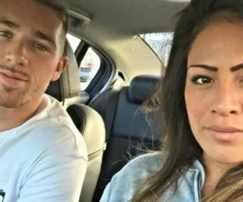 Two people are sitting in a car, looking towards the camera.