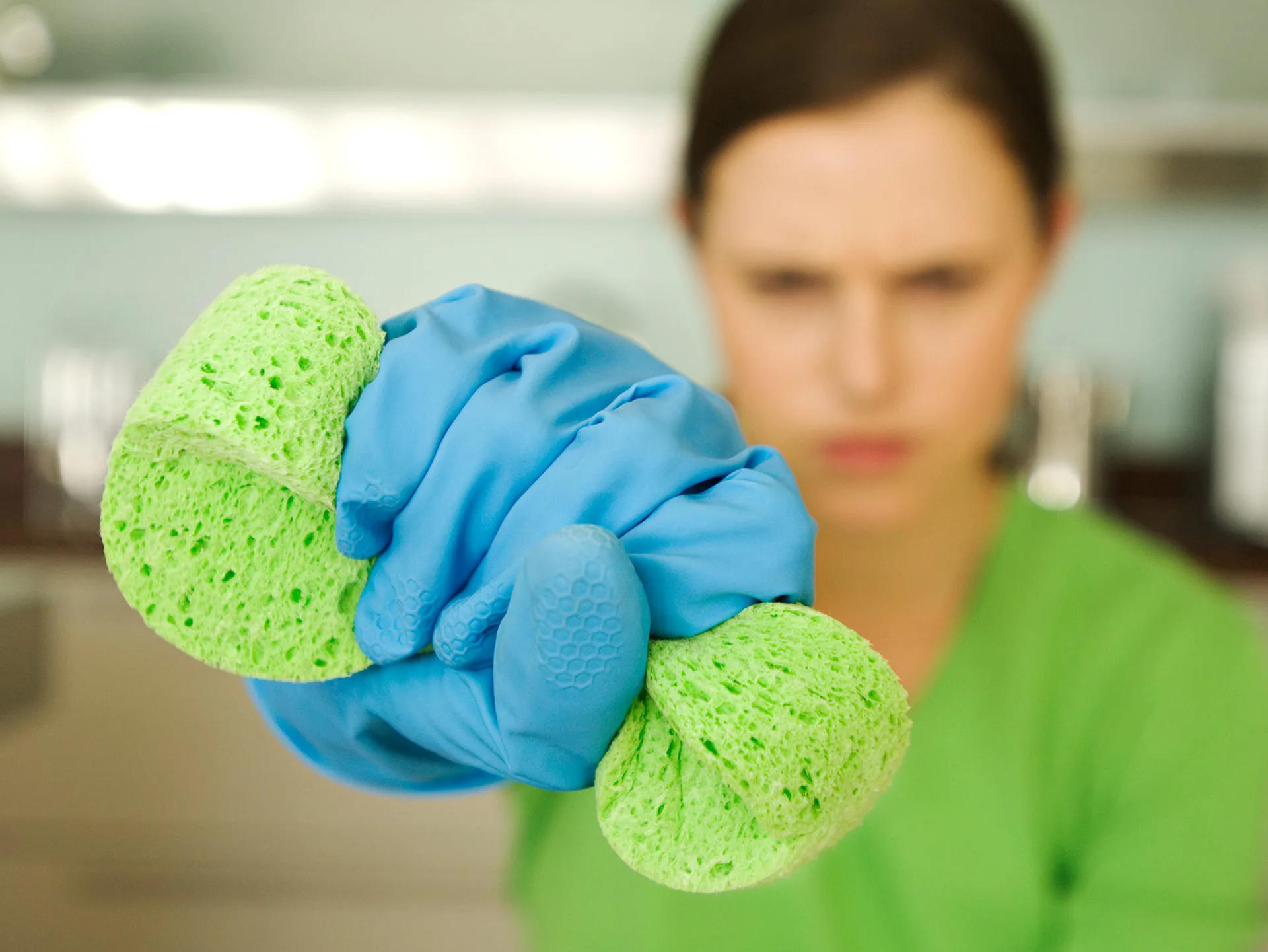 A person in a green shirt and blue gloves holds a green sponge in a kitchen setting.